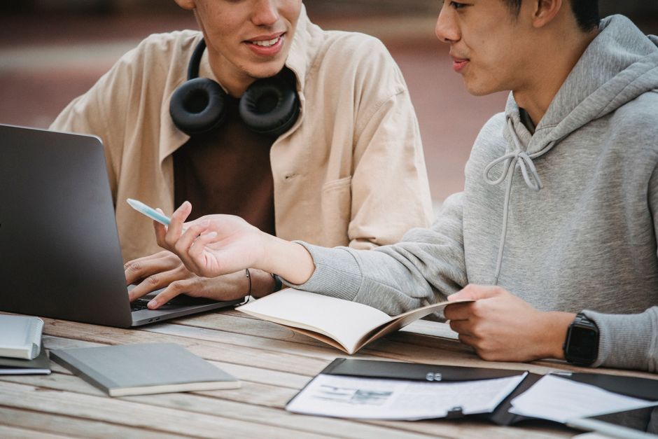 Crop anonymous multiethnic male students in casual clothes at table with notebook and laptop working on assignment together