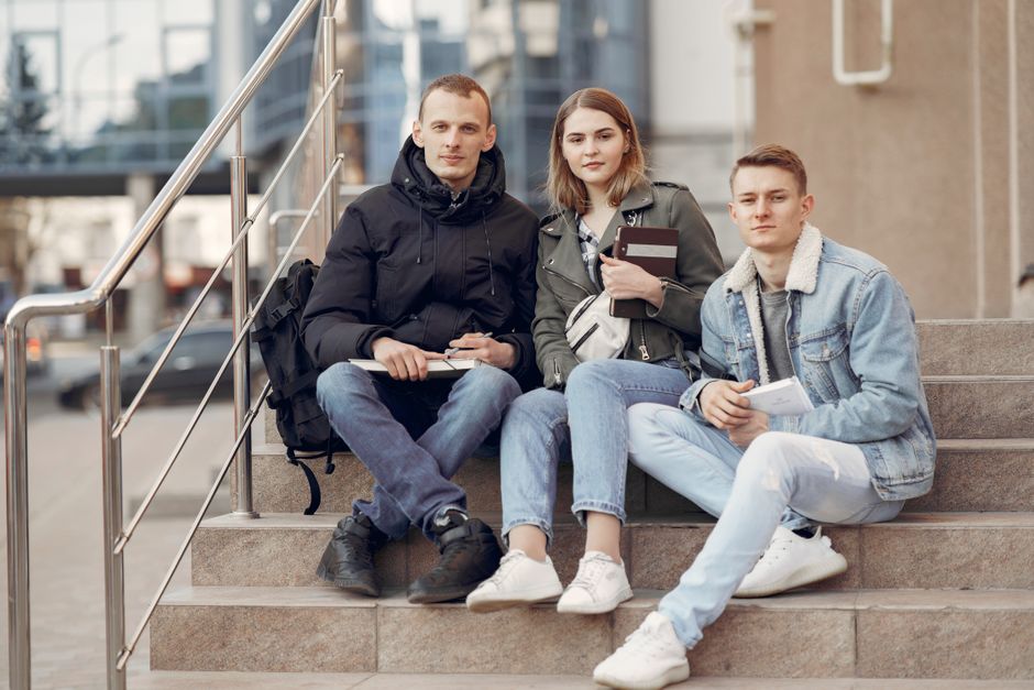 Group of young students in casual wear and sneakers sitting on stairs with books near modern building on street in daylight and looking at camera